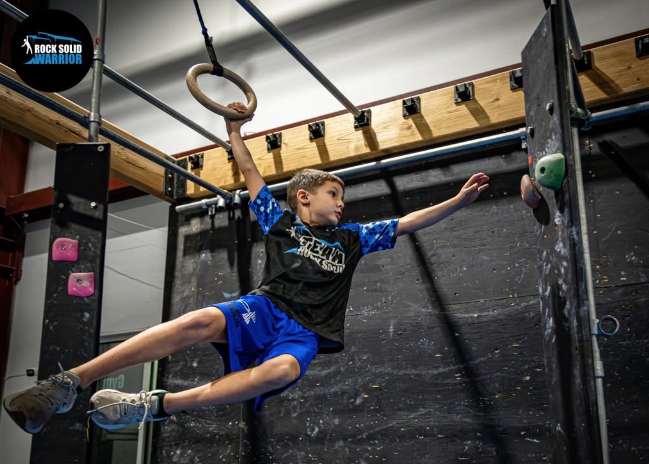 Young boy mid-air on a ninja warrior obstacle course, gripping a hanging ring with one hand while reaching for a climbing hold with the other, at Rock Solid Warrior Apex gym.